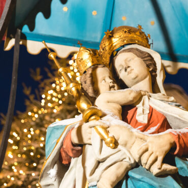 Das Foto zeigt eine farbenfrohe Statue der Madonna mit dem Jesuskind, beide mit goldenen Kronen geschmückt. Maria hält das Kind liebevoll im Arm, während sie einen goldenen Zepter trägt. Die Statue steht unter einem blauen Baldachin, im Hintergrund sind Lichter eines Weihnachtsbaums zu sehen.