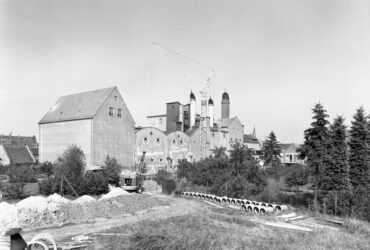 Das Foto zeigt die Hochheimer Malzfabrik während einer Bauphase, bei der ein Kran und Baumaschinen im Vordergrund zu sehen sind. Die markanten Schornsteine und das Fabrikgebäude prägen das Stadtbild, das durch die Malzfabrik über Jahrzehnte hinweg entscheidend beeinflusst wurde.