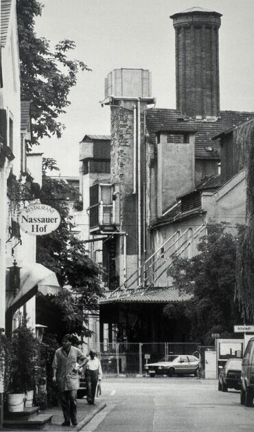 Das Bild zeigt die Hochheimer Malzfabrik im Hintergrund der Altstadt, mit ihrem markanten Schornstein und den umliegenden industriellen Bauten. Im Vordergrund befindet sich das Restaurant "Nassauer Hof", und die Szene vermittelt eine kontrastreiche Mischung aus historischer Architektur und industrieller Umgebung.