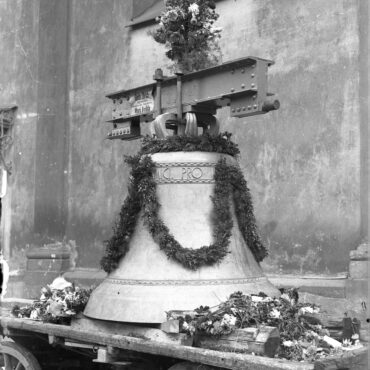 Eine große Kirchenglocke, geschmückt mit Blumen und Girlanden, steht auf einem hölzernen Wagen vor einer Kirche. Über der Glocke ist eine Metallhalterung befestigt, und im Hintergrund ist eine Wand mit einem Kirchenfenster zu sehen.