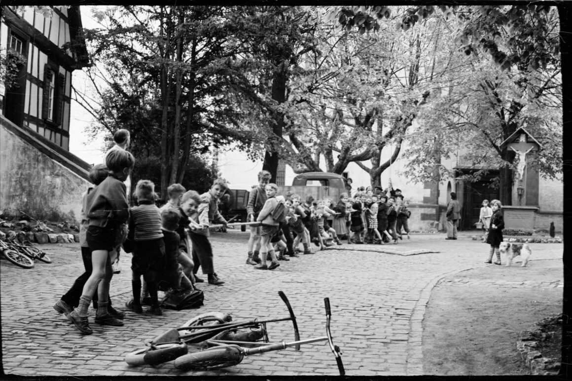 Eine große Gruppe von Kindern und Erwachsenen nimmt an einem Seilziehen vor der katholischen Kirche teil. Im Hintergrund ist die Kirche mit einem Kreuz zu sehen