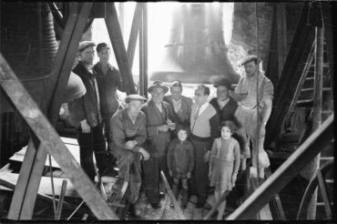 Eine Gruppe von Menschen, darunter Männer, Frauen und Kinder, posiert vor einer großen Glocke im Turm der katholischen Kirche.