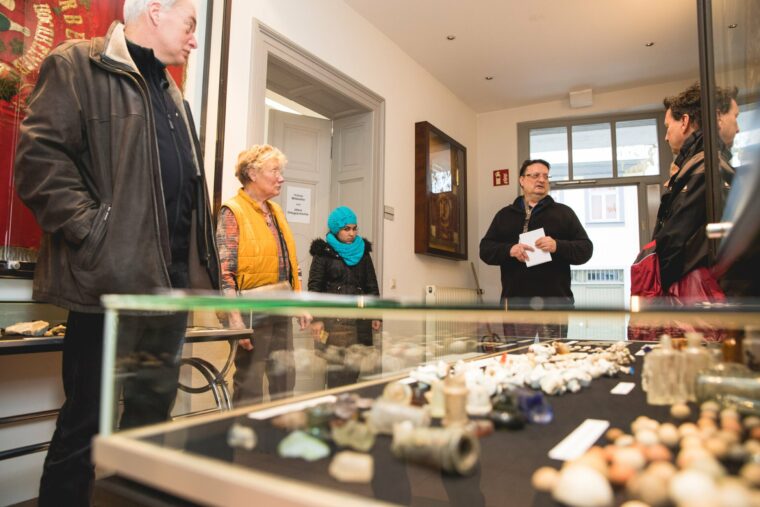 Auf dem Bild ist eine Gruppe von Menschen im Ausstellungsraum des Hochheimer Heimatmuseums zu sehen. Ein Mann spricht zu den Besuchern, während sie um eine Vitrine mit verschiedenen historischen Artefakten stehen. Die Atmosphäre wirkt konzentriert und interessiert, während die Gruppe den Erklärungen lauscht.