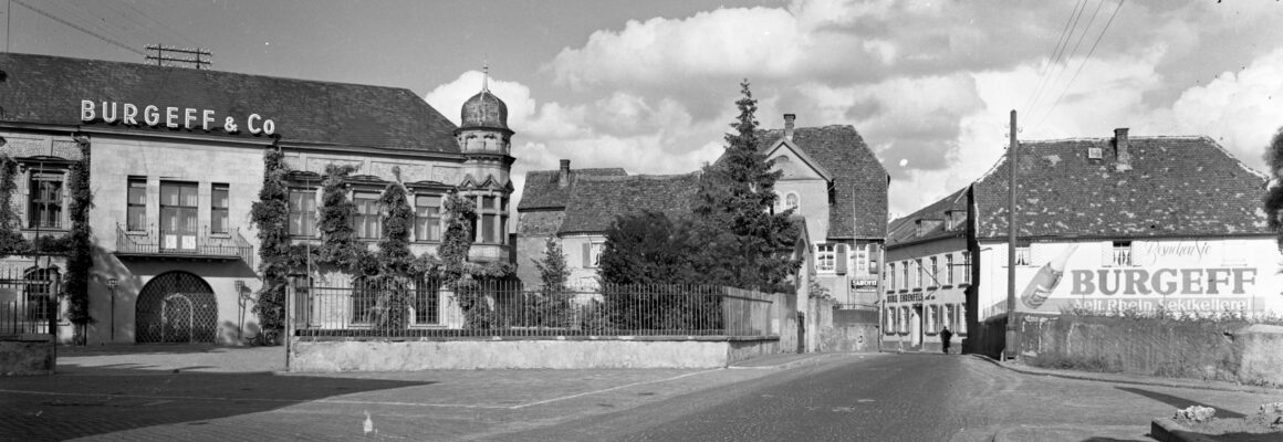 Ein großes historisches Gebäude mit der Aufschrift "Burgeff & Co" auf dem Dach. Die Fassade ist von Efeu bewachsen, und die Straße führt an der Anlage vorbei.