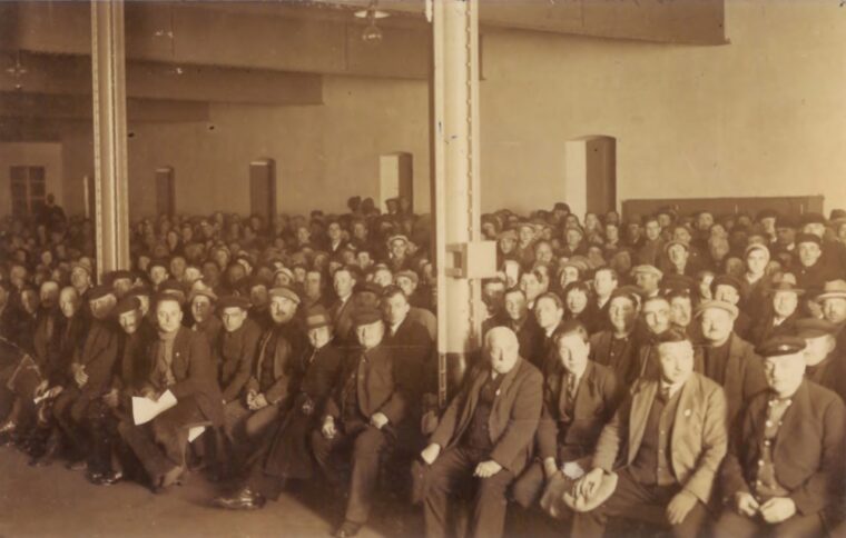 Das Bild zeigt eine große Gruppe von Menschen, die in der Zepplinhalle sitzen. Die Personen tragen überwiegend Hüte und Anzüge, und das Foto hat einen sepiafarbenen Ton, der ihm ein historisches Aussehen verleiht. Zwei Stützsäulen teilen den Raum, was auf eine geräumige Halle hindeutet.