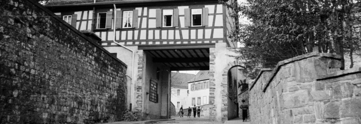 Das Foto zeigt das Küsterhaus mit Stadttor, das über einer steinernen Durchfahrt steht. Der Zugang führt durch ein Tor, das in den Stadtkern von Hochheim führt.