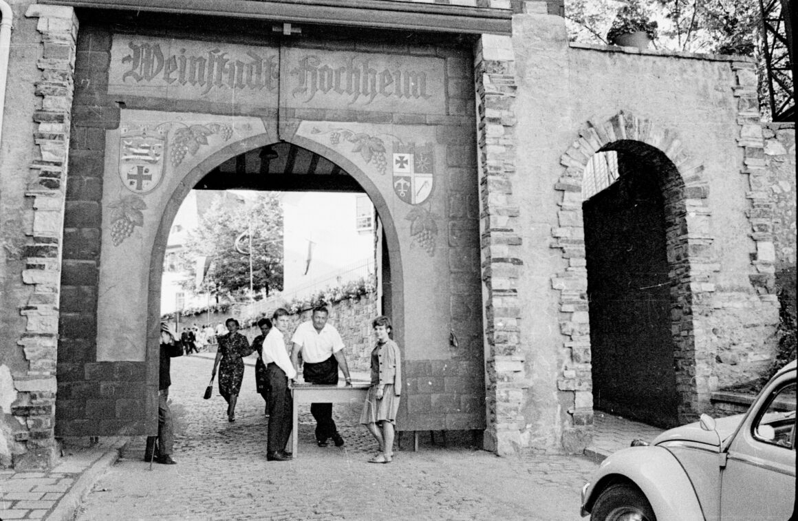 Das Foto zeigt das Weinfest in Hochheim im Jahr 1963, bei dem Kassierer der Vereine am Tor des Küsterhauses die ankommenden Besucher begrüßen. Das historische Stadttor ist festlich dekoriert, und die Besucher benötigen ein Festabzeichen, um Zutritt zum Fest zu erhalten. Im Vordergrund parkt ein VW Käfer, während im Hintergrund weitere Festbesucher die gepflasterte Straße entlanggehen.