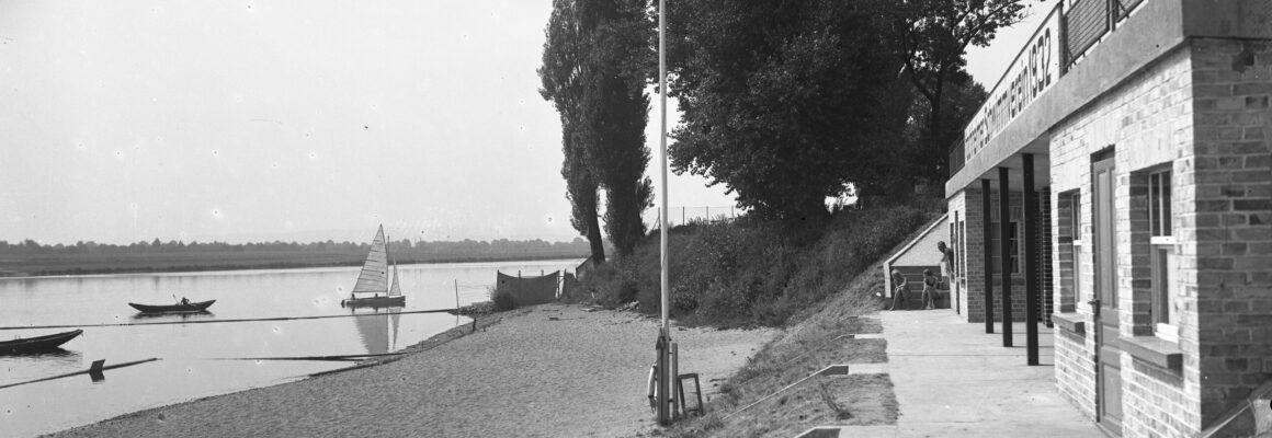 Das Foto zeigt das Hochheimer Strandbad am Main. Im Wasser befindet sich ein Segelboot. Rechts im Bild steht das Gebäude vom Strandbad, in denen sich Umkleiden befinden.