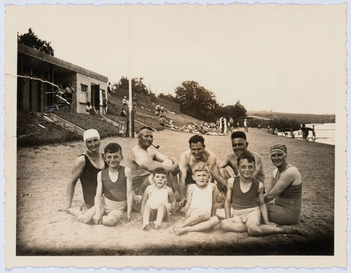 Das Bild zeigt eine Gruppe von Menschen, die beim Strandbad am Main auf dem Sand sitzt. Die Gruppe besteht aus Männern, Frauen und Kindern, die in Badeanzügen gekleidet sind, während im Hintergrund weitere Personen am Ufer des Flusses entspannen. Die Atmosphäre wirkt fröhlich und entspannt, vermutlich handelt es sich um einen Familienausflug oder einen gemeinsamen Tag am Strand.