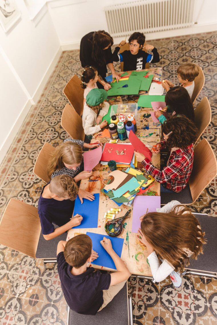 Eine Gruppe von Kindern sitzt an einem Tisch in einem Kunstunterricht und arbeitet an bunten Bastelprojekten. Eine Betreuerin hilft einem Kind, während die anderen konzentriert auf ihre Werke blicken.