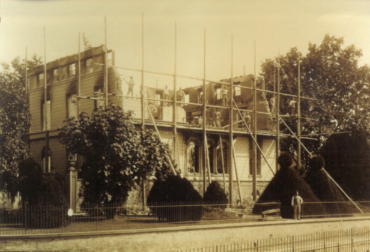 Das Bild zeigt den Wiederaufbau der Villa Burgeff, die von einem Gerüst umgeben ist. Mehrere Arbeiter sind auf dem Gerüst zu sehen, während sie an der Struktur arbeiten. Vor dem Gebäude befindet sich ein gepflegter Garten mit Hecken, die kunstvoll geschnitten sind, und ein Mann steht an einem Zaun vor der Villa.