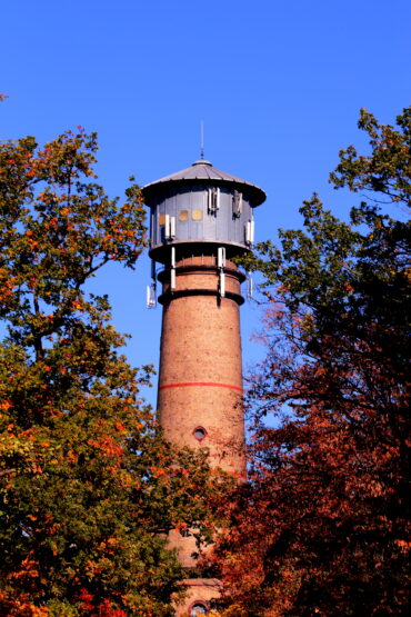 Der Hochheimer Wasserturm erhebt sich über die herbstlich gefärbten Baumkronen und sticht mit seinem markanten Ziegelmauerwerk und dem runden Wassertank ins Auge. Das klare Blau des Himmels bildet einen beeindruckenden Kontrast zu den warmen Herbstfarben, die den Turm umrahmen.