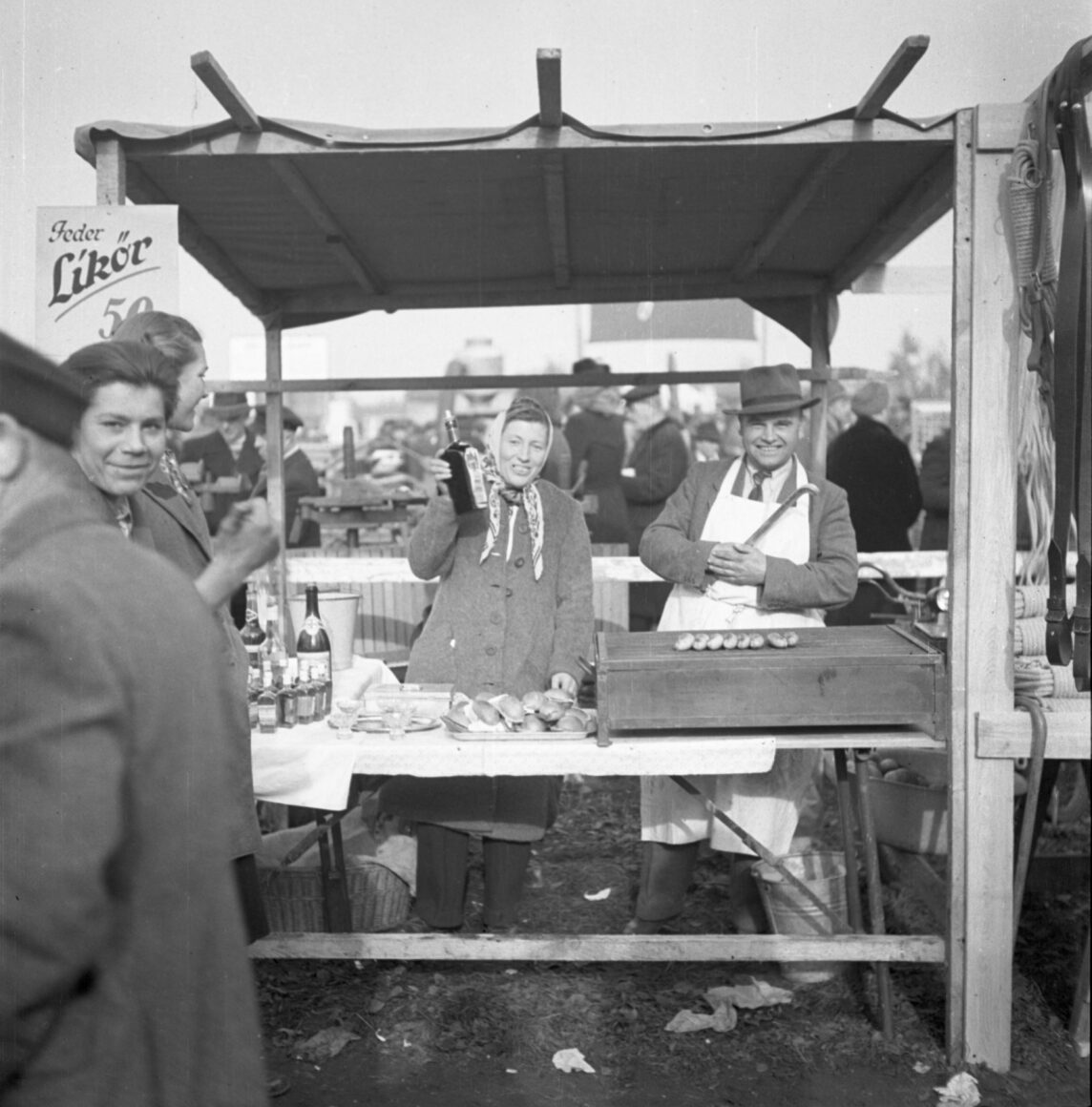 Das Bild zeigt einen Verkaufsstand auf dem Hochheimer Markt, an dem Likör und Speisen angeboten werden. Eine Frau in einem Mantel und Kopftuch präsentiert lächelnd eine Flasche Likör, während ein Mann im Hintergrund an einem Grill Würstchen zubereitet. Um den Stand herum stehen weitere Personen, die sich die Produkte anschauen.