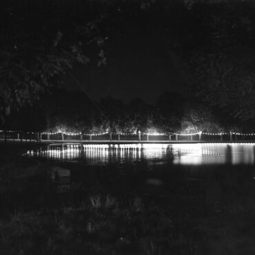 Das Bild zeigt eine nächtliche Szenerie am Hochheimer Weiher, der von Lichterketten beleuchtet wird. Die Lichter reflektieren im Wasser und sorgen für eine stimmungsvolle Atmosphäre. Umgeben von Bäumen, ist die Szenerie ruhig und idyllisch.