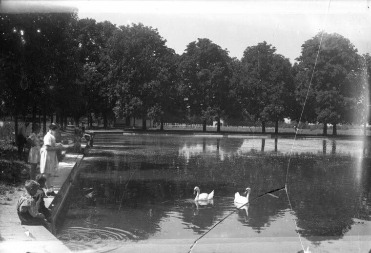 Das Bild zeigt den Hochheimer Weiher um das Jahr 1933. Am Ufer des Weihers stehen mehrere Personen, darunter Kinder und Erwachsene, die das ruhige Gewässer mit zwei schwimmenden Schwänen betrachten. Im Hintergrund sind große Bäume zu sehen, die den Weiher umgeben und eine friedliche, idyllische Atmosphäre schaffen.