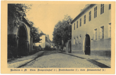 Das Bild zeigt eine gepflasterte Straßenansicht in Hochheim. Auf der rechten Seite befindet sich der ehemalige Dompräsenzhof, während auf der linken Seite das Domdechanetor zu sehen ist. Im Hintergrund ist der ehemalige Johanniterhof sichtbar. Die Gebäude zeichnen sich durch historische Architektur mit Torbögen und Fenstern mit Fensterläden aus, eingebettet in eine von Bäumen gesäumte Straße, die einen ruhigen und charmanten Eindruck vermittelt.