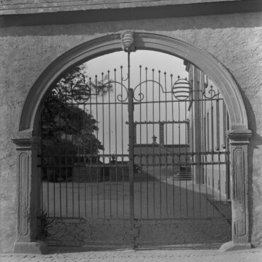 Das Bild zeigt das Tor des Domänenhofs, ein kunstvoll geschmiedetes Eisentor in einem breiten Rundbogen. Über dem Torbogen ist ein dekoratives Element angebracht, das dem Tor zusätzlichen Charakter verleiht. Im Hintergrund ist ein Teil des Domänenhofs zu sehen.