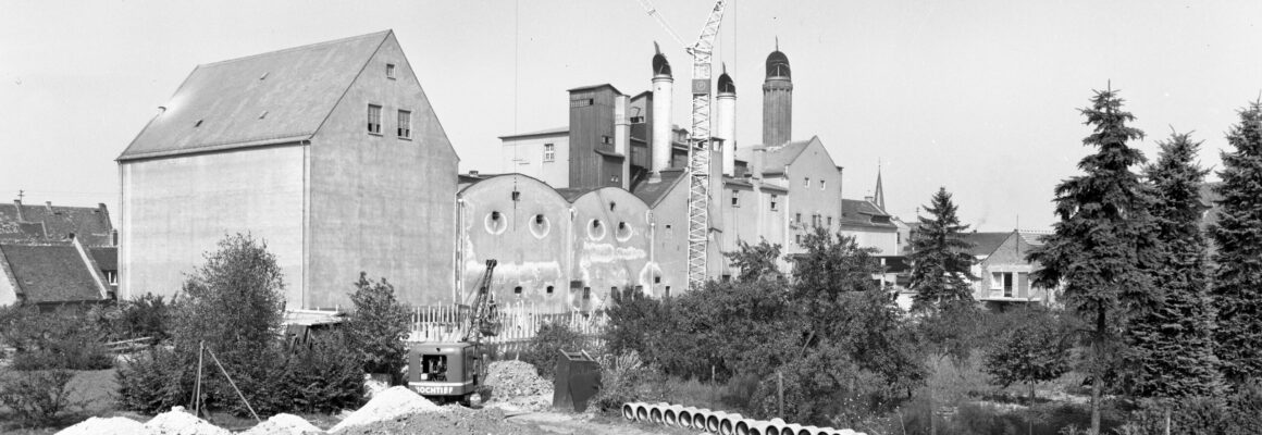 Eine Fabrikanlage mit mehreren Gebäuden und einem Baukran. Vor der Fabrik sind Baustellenfahrzeuge und Rohre sichtbar, während Bäume das Gelände umgeben.