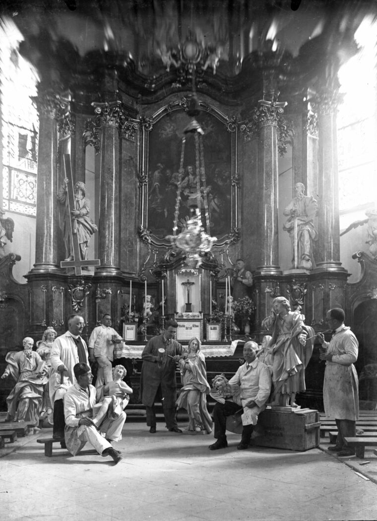 Das Bild zeigt eine Gruppe von Restauratoren in der Pfarrkirche St. Peter und Paul in Hochheim. Sie arbeiten an verschiedenen religiösen Skulpturen und Figuren, die um den Altar herum aufgestellt sind. Im Hintergrund ist der prunkvolle Hochaltar der Kirche zu sehen, geschmückt mit religiösen Darstellungen und kunstvollen Verzierungen, während die Restauratoren konzentriert bei ihrer Arbeit sind.