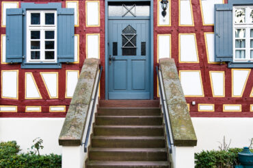 Das Bild zeigt den Eingang des Küsterhauses in Hochheim, ein historisches Fachwerkhaus mit blauen Fensterläden und einer robusten Steintreppe. Die Fassade ist mit rot-braunen Balken und gelben Akzenten geschmückt, die das Fachwerk betonen.