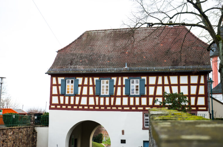 Das Bild zeigt eine Gesamtansicht des Küsterhauses mit dem darunterliegenden Stadttor. Das Fachwerkhaus hat eine symmetrische Anordnung der Fenster mit blauen Fensterläden und ein charakteristisches Dach, das die historische Architektur der Region widerspiegelt. Der Torbogen unter dem Gebäude führt zu einem Weg und fügt sich harmonisch in die Umgebung ein.