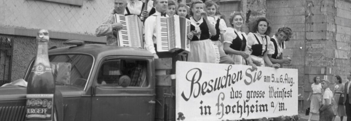 Das Bild zeigt eine Gruppe von Musikern und Frauen in traditioneller Tracht auf einem geschmückten Wagen, der Teil des Hochheimer Weinfestes von 1950 ist. Auf dem Wagen ist ein großes Schild angebracht, das die Besucher zum Weinfest am 5. und 6. August in Hochheim am Main einlädt. Eine übergroße Nachbildung einer Sektflasche der Marke Burgeff ist ebenfalls auf dem Wagen prominent platziert.